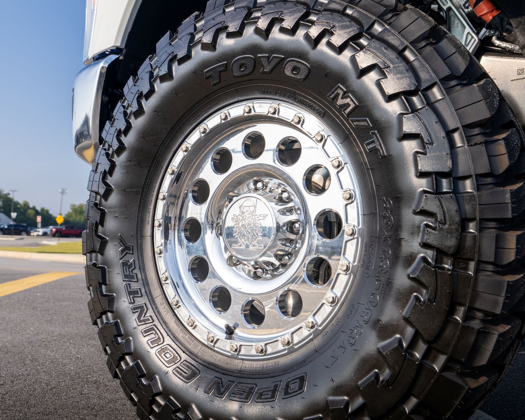 A close-up shot of a black Toyo MT / DTN All Terrain tire mounted on a polished Custom Wild Willies silver multi-spoke wheel, attached to a white vehicle on an asphalt road.